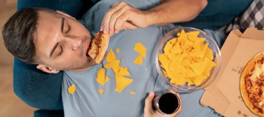 homem deitado comendo pizza, com nachos caídos no colo e segurando um copo de refrigerante