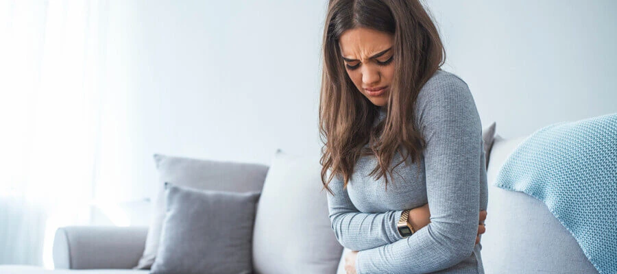 Mulher sentada no sofá com as mãos na barriga e expressão de dor